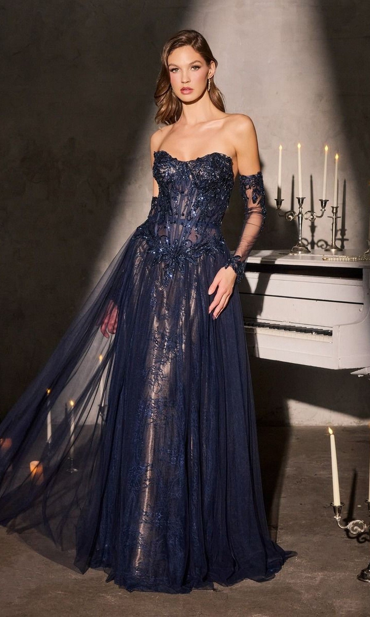 A woman in a strapless black beaded prom gown (Ladivine CD717) stands beside a white piano topped with glowing candelabras in a dimly lit room.